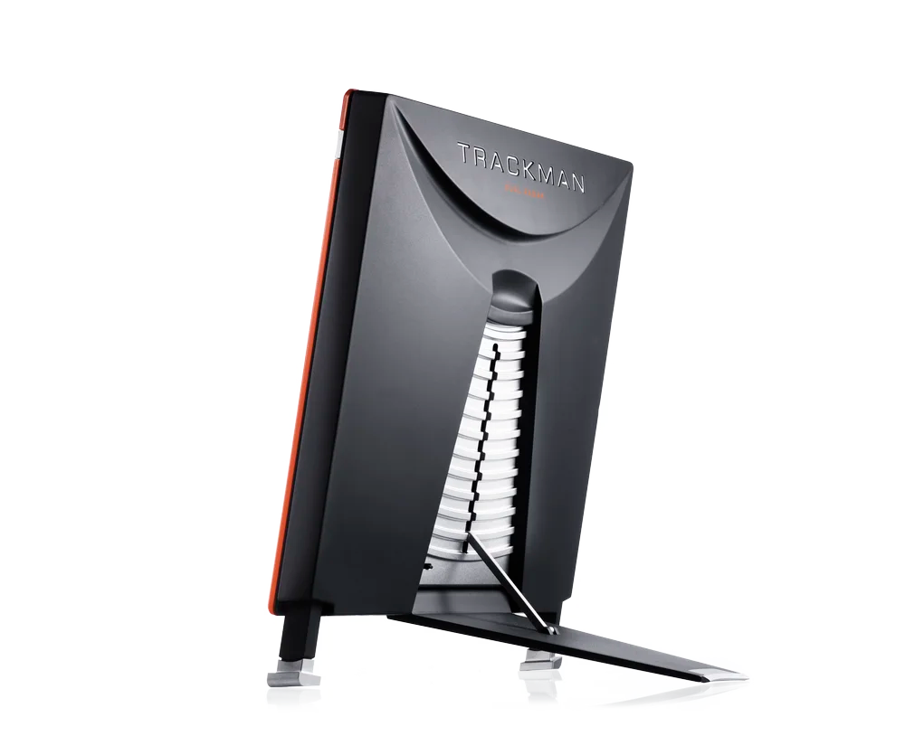 L'image montre un appareil de forme carrée, compact, avec une face avant orange portant l'inscription "TrackMan". Sur la vue latérale, on remarque un pied de support inclinable pour la stabilisation de l'appareil. La vue arrière montre un design noir avec des fentes d'aération et un système radar intégré, avec l'inscription "TrackMan" en haut et la mention "Dual Radar" en dessous.
